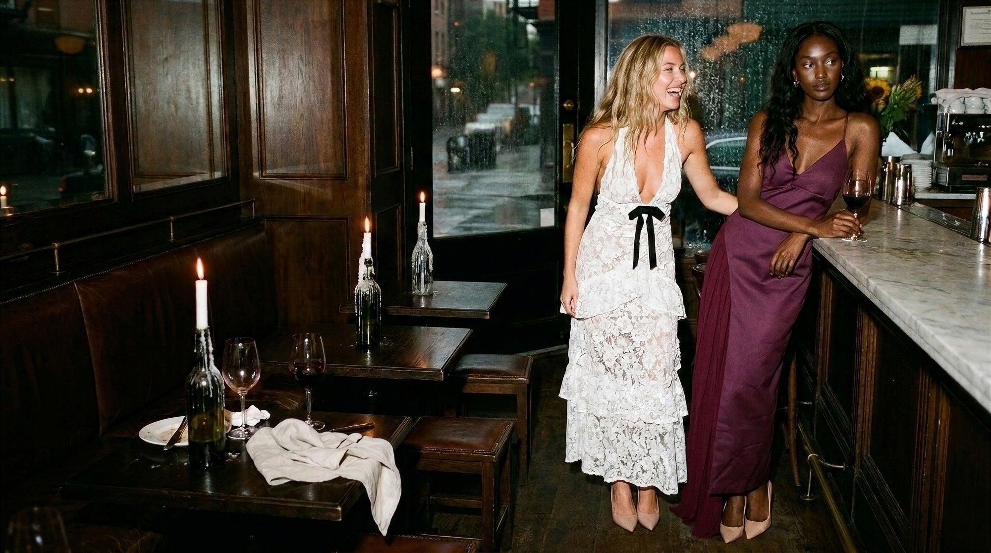 Two friends in a white lace tiered maxi dress and a wine satin slip maxi dress at a candlelit restaurant bar on a rainy autumn night